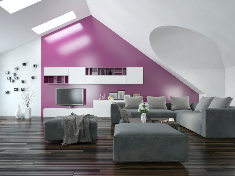 Modern apartment living room interior with a purple accent wall and sloping ceiling with skylights above a parquet floor and modern grey lounge suite with wall cabinets and television