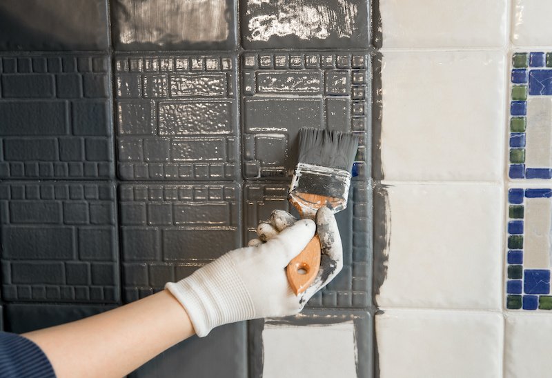 Repainting old dated kitchen ceramic tile back wall with modern gray chalk paint indoors at home. Giving old kitchen new look concept. Hand holding a paint brush tool.