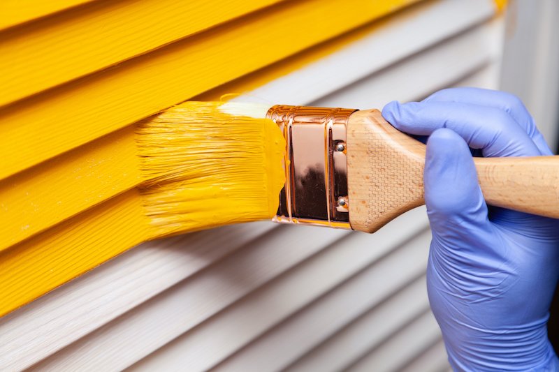 Closeup woman hand in purple rubber glove with paintbrush painting natural wooden door with yellow paint. Concept creative design house interior. How to Paint Wooden Surface. Selected focus