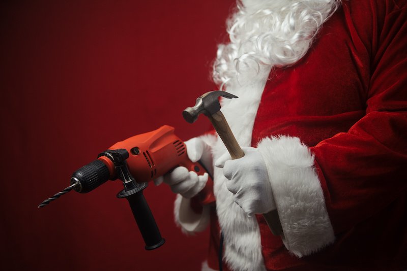 Father Santa Claus holding using electric drill and hammer tools ready to renovate space background. Handyman repairman in festive mood for seasonal business work. Merry Christmas and Happy New Year!
