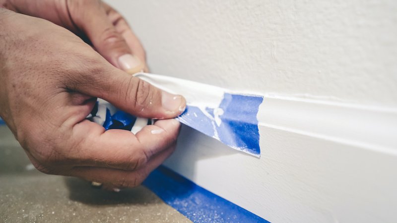 Removing masking tape from molding. A painter pulls of blue painter's tape from the wall to reveal a clean edge baseboard.
