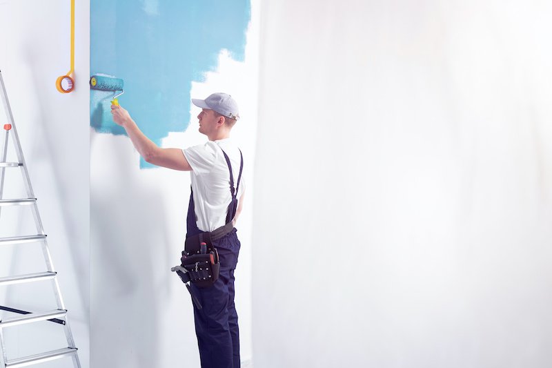 Decorator wearing a overall painting a room on a blue color. Place your graphic/tool/logo on the empty wall