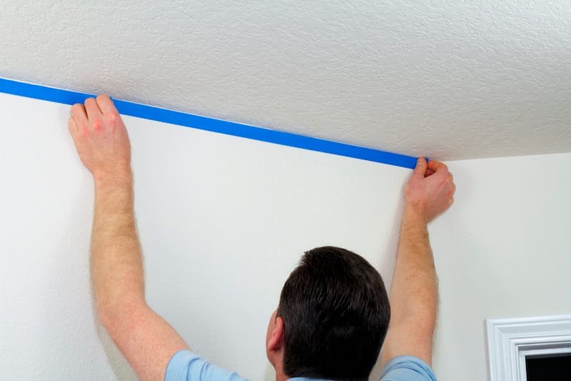 Man preparing to paint ceiling by masking off the wall beneath it with blue painter's tape.