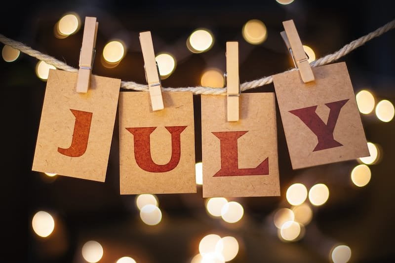 The word JULY printed on clothespin clipped cards in front of defocused glowing lights.