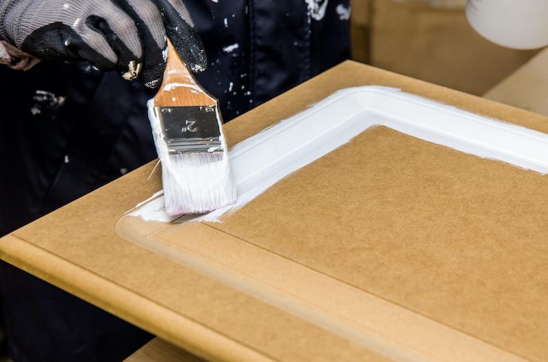 Repainting kitchen cabinet doors with white chalk paint indoors at home. Giving old kitchen new look concept. Hand holding a paint brush tool with paint against old cupboard door.